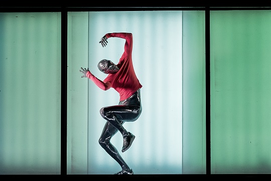 A dancer performing in a full body latex suit, with the inside of a green truck behind them.