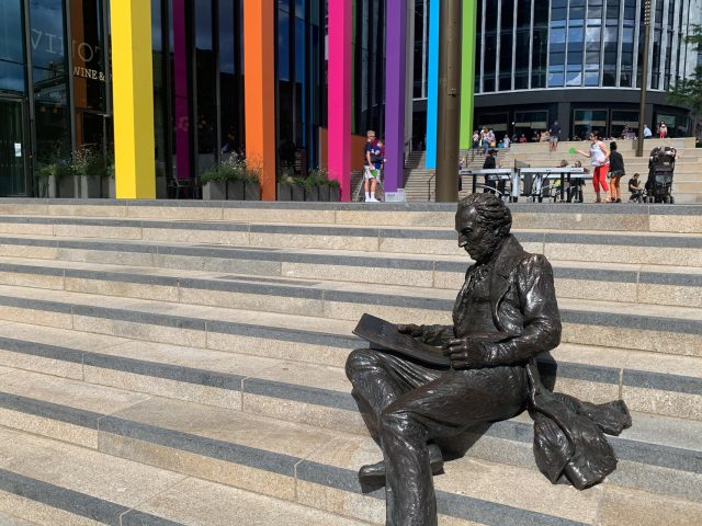 Thomas Attwood statue in Chamberlain Square, Paradise Birmingham. The columns of Two Chamberlain Square are painted in the Commonwealth Games colours behind him.