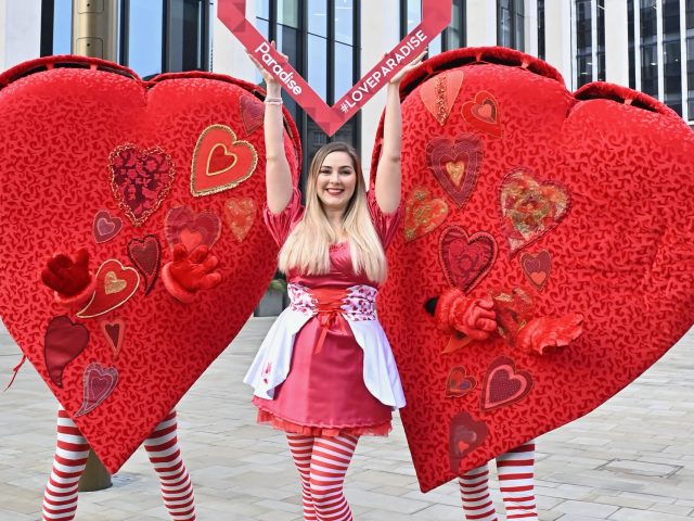 Woman dressed in a red dress, with people in love heart costumes on either side of her, holding a heart picture frame.